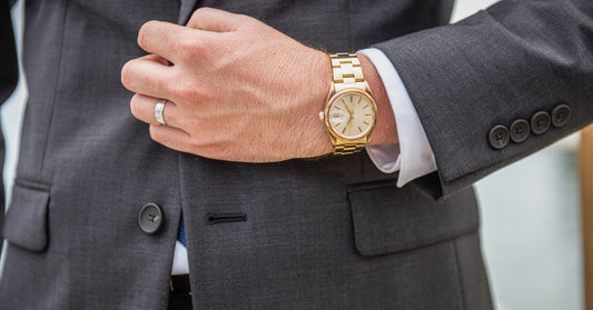 A man wearing a luxury gold watch uses his left hand to hold his suit jacket closed. The golden watch bracelet glows.