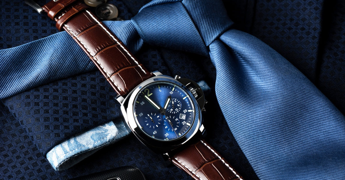 Close-up of a luxury watch on top of a blue suit and tie. The watch has a brown leather strap and a dark blue dial.