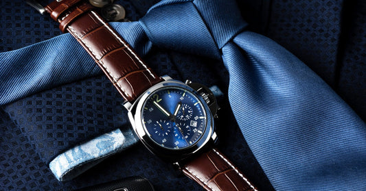 Close-up of a luxury watch on top of a blue suit and tie. The watch has a brown leather strap and a dark blue dial.