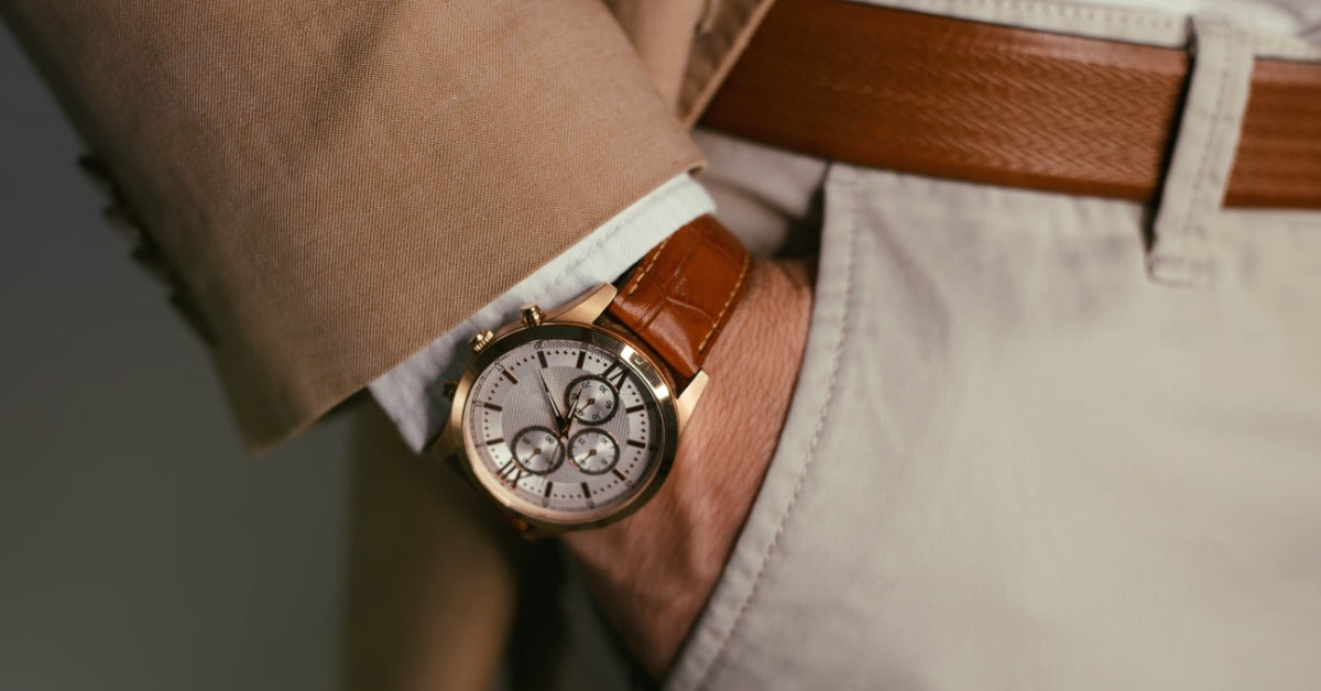 A watch with a white face, subdials, and a brown leather strap is on a man’s wrist as his hand rests in his pocket.