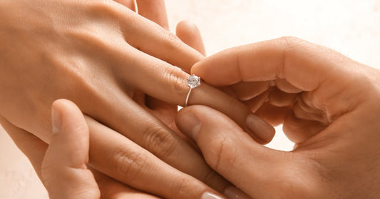 A close-up of one person holding another person's manicured hand and placing a diamond ring on their ring finger.