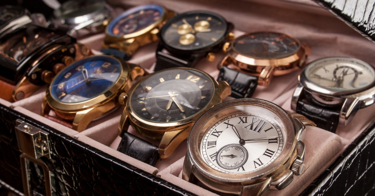 A watch storage case with a light pink interior holds analog watches in different colors, metals, and dial styles.